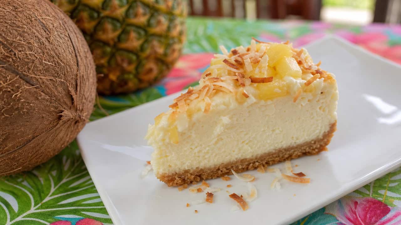 a pineapple coconut cheesecake in a wide horizontal shot, creamy slice topped with toasted coconut and pineapple on a coconut crust, on a white plate, on a bright tropical-themed table, natural light, shallow depth of field, landscape orientation