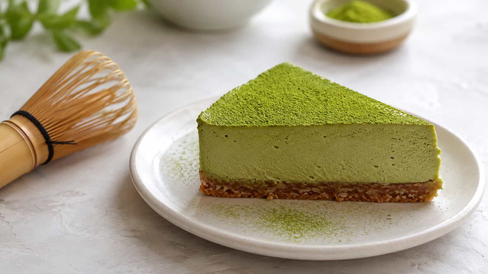 a matcha green tea cheesecake in a wide horizontal shot, vivid green slice dusted with matcha on a coconut crust, on a white plate beside a bamboo whisk, on a light table, natural light, shallow depth of field, landscape orientation