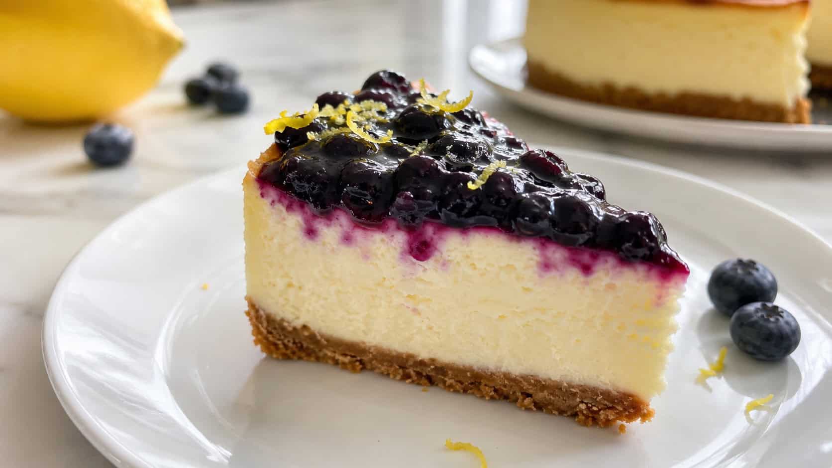 a lemon blueberry cheesecake in a wide horizontal shot, pale yellow slice topped with glossy blueberry compote and lemon zest, on a white plate, on a bright counter, natural light, shallow depth of field, landscape orientation