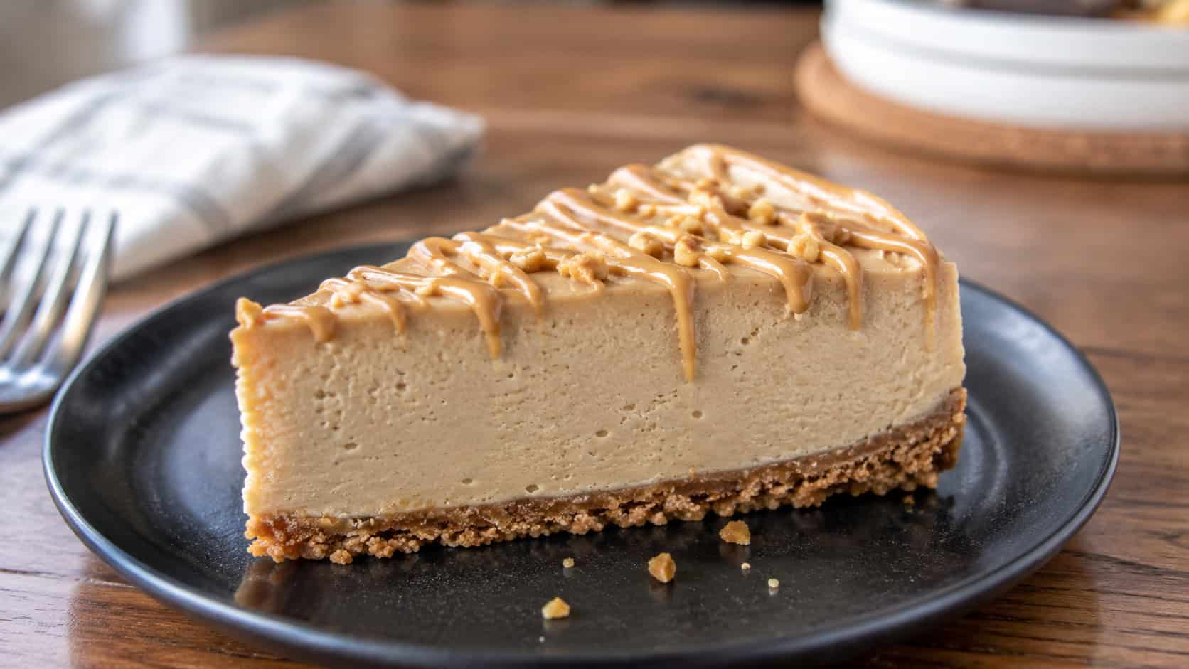a baked peanut butter cheesecake in a wide horizontal shot, thick tan slice with a peanut butter drizzle on a graham crust, on a dark plate, on a wooden table, natural light, shallow depth of field, landscape orientation