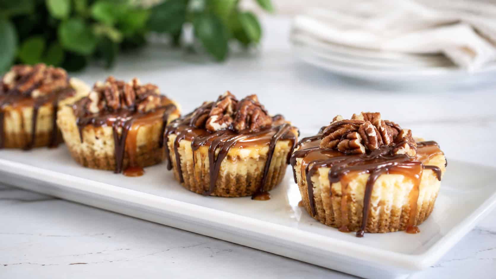 mini turtle cheesecake tarts in golden shells topped with caramel, chocolate drizzle, and pecans on a white platter, in a wide horizontal shot, arranged in a row, on a bright table, natural light, shallow depth of field, landscape orientation