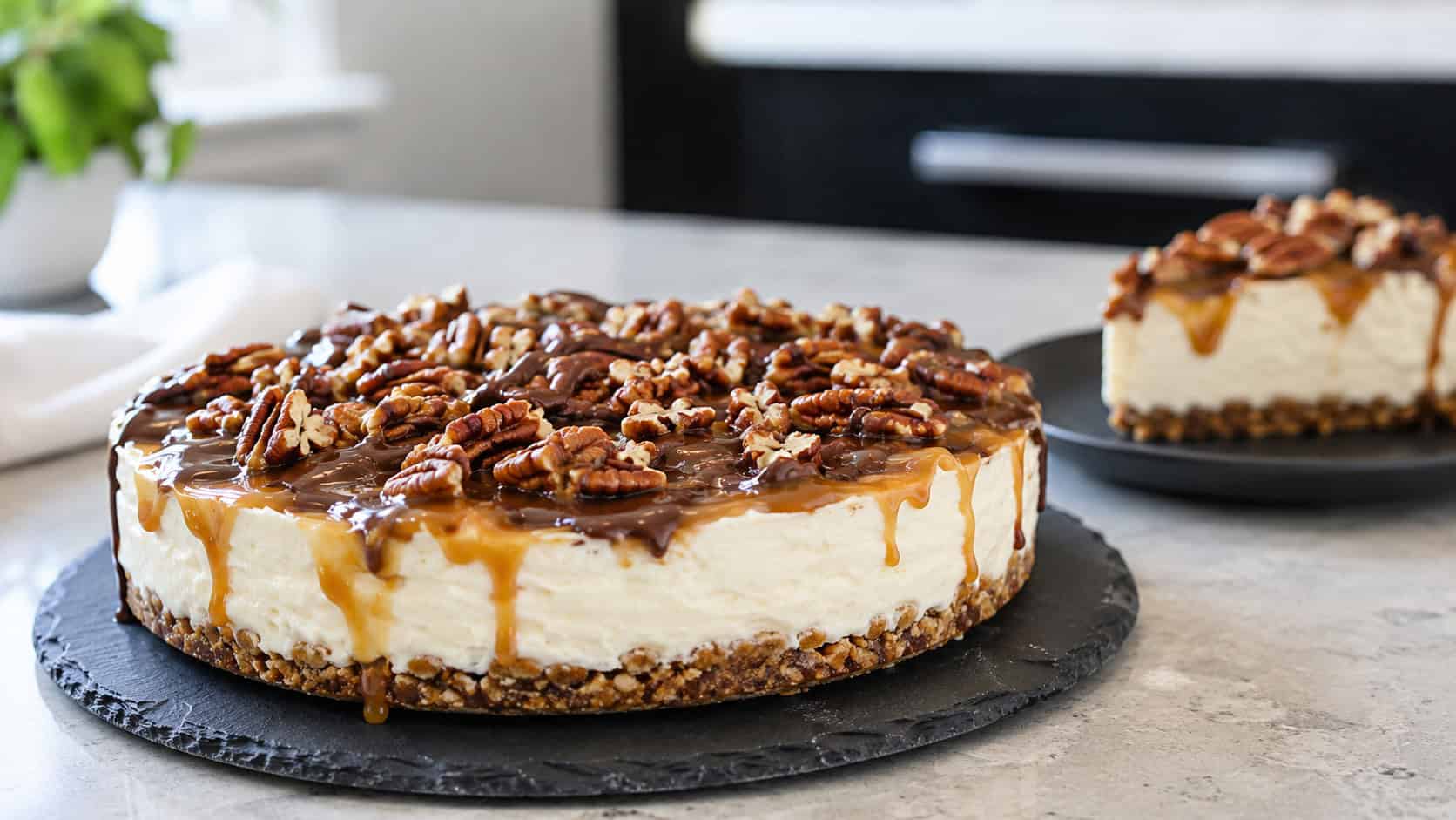 a keto no-bake turtle cheesecake with an almond crust topped with sugar-free caramel, chocolate, and pecans on a slate plate, in a wide horizontal shot, a slice on the side, on a modern counter, natural light, shallow depth of field, landscape orientation