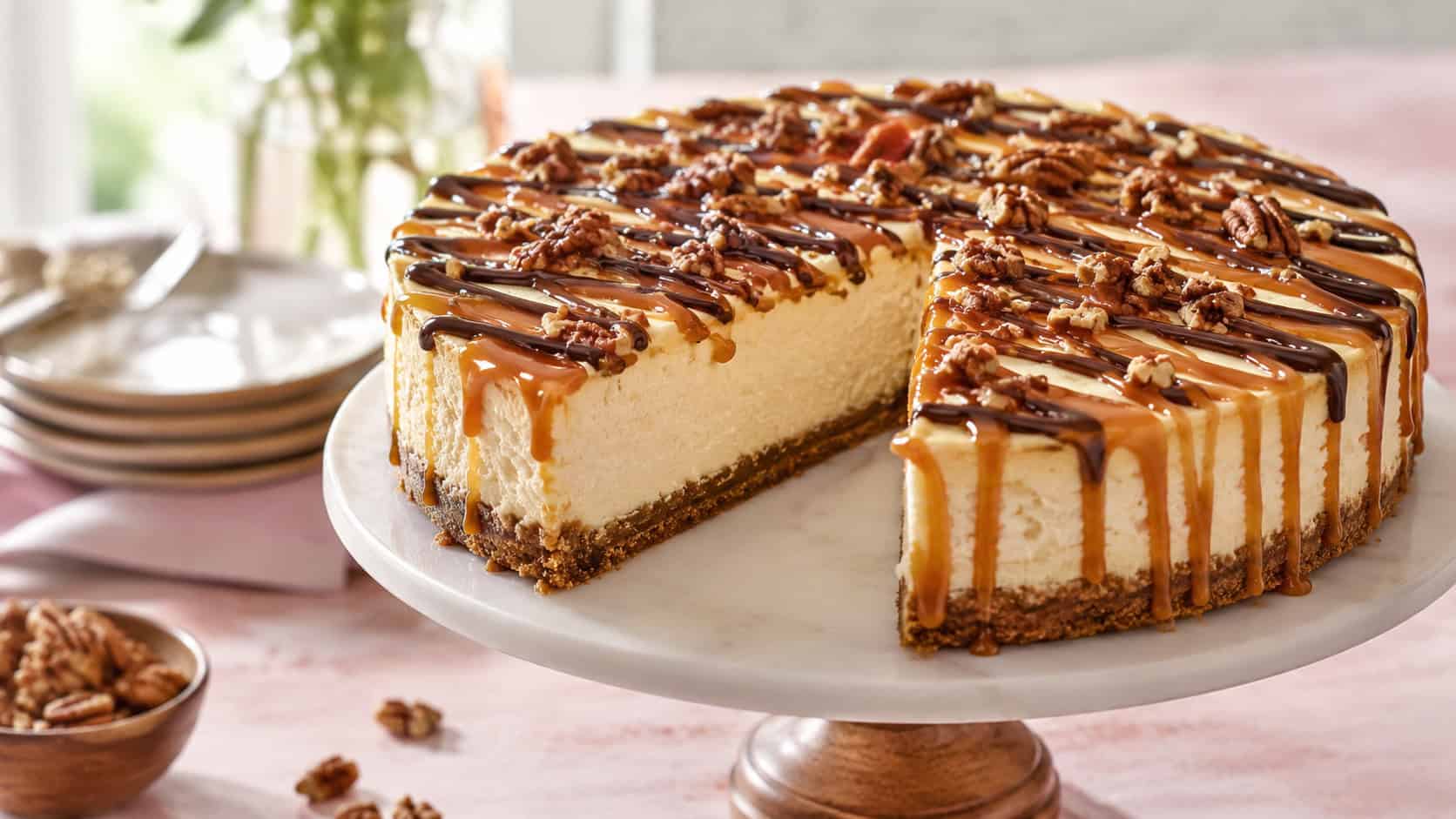a pale creamy white chocolate turtle cheesecake topped with caramel, dark chocolate drizzle, and pecans on a cake stand, in a wide horizontal shot, a slice removed, on a soft pastel table, natural light, shallow depth of field, landscape orientation