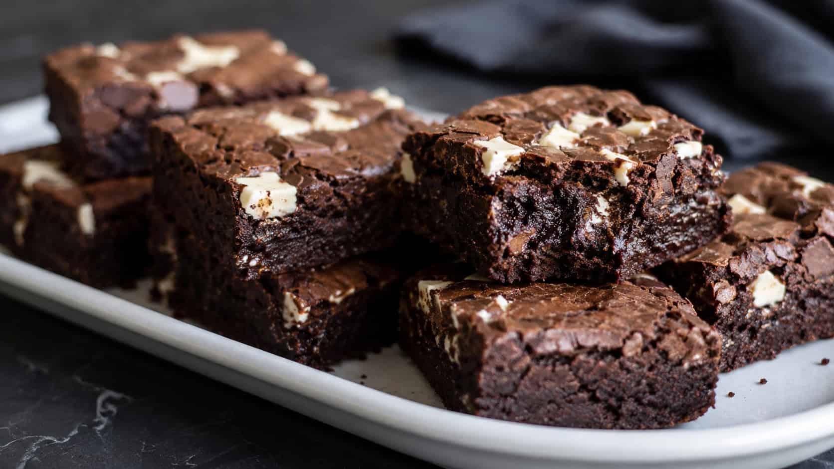 a wide horizontal platter of dense triple chocolate brownies with dark and white chocolate chunks, one broken to show a fudgy truffle interior, on a dark surface, moody natural light, shallow depth of field, landscape orientation