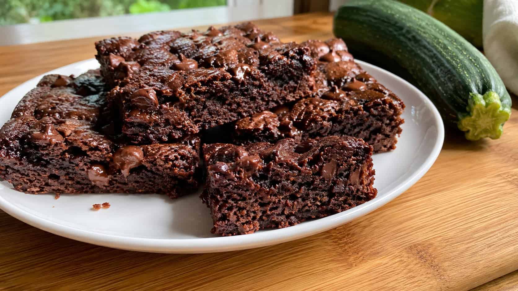 a wide horizontal plate of moist chocolate brownies with melting chips, one piece showing a tender crumb, on a wooden board with a fresh zucchini nearby, natural light, shallow depth of field, landscape orientation