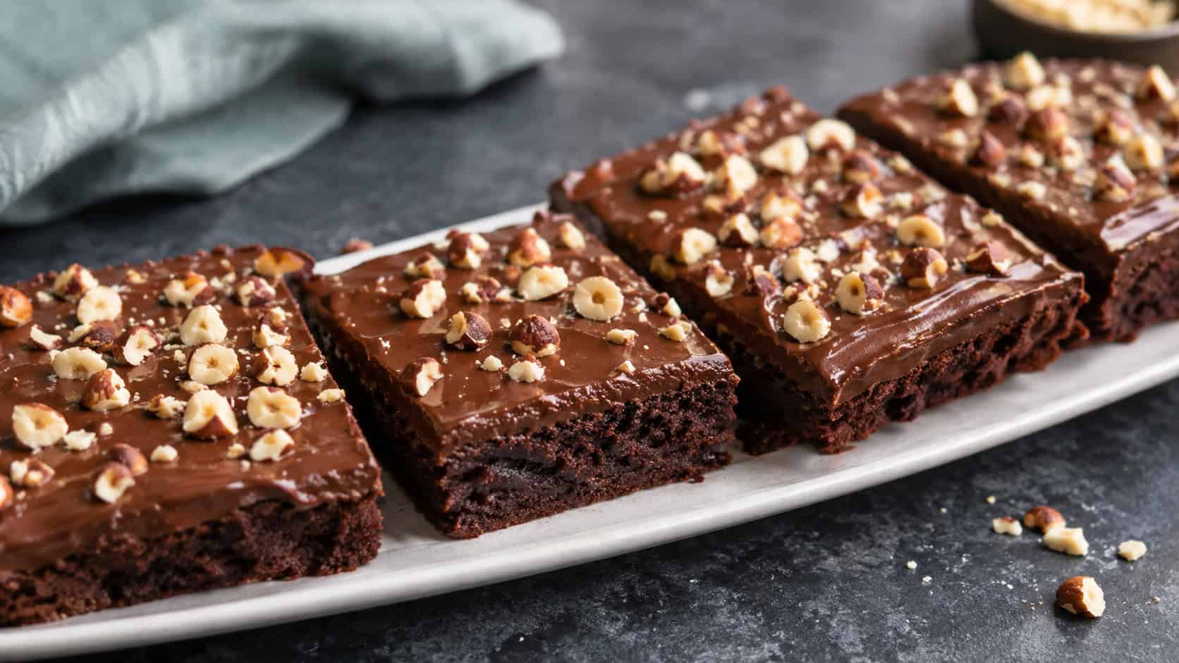 a wide horizontal platter of brownies topped with glossy chocolate ganache and chopped hazelnuts, one cut to show the fudgy base, on a dark surface, soft natural light, shallow depth of field, landscape orientation