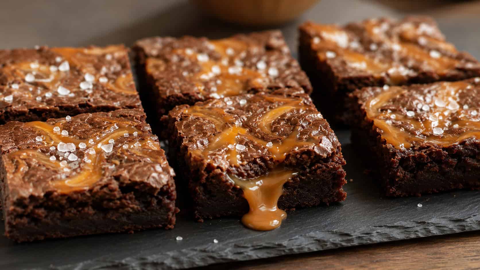 a wide horizontal arrangement of chocolate brownies with golden caramel swirls and flaky salt on top, one piece with caramel oozing out, on a dark slate board, warm natural light, shallow depth of field, landscape orientation