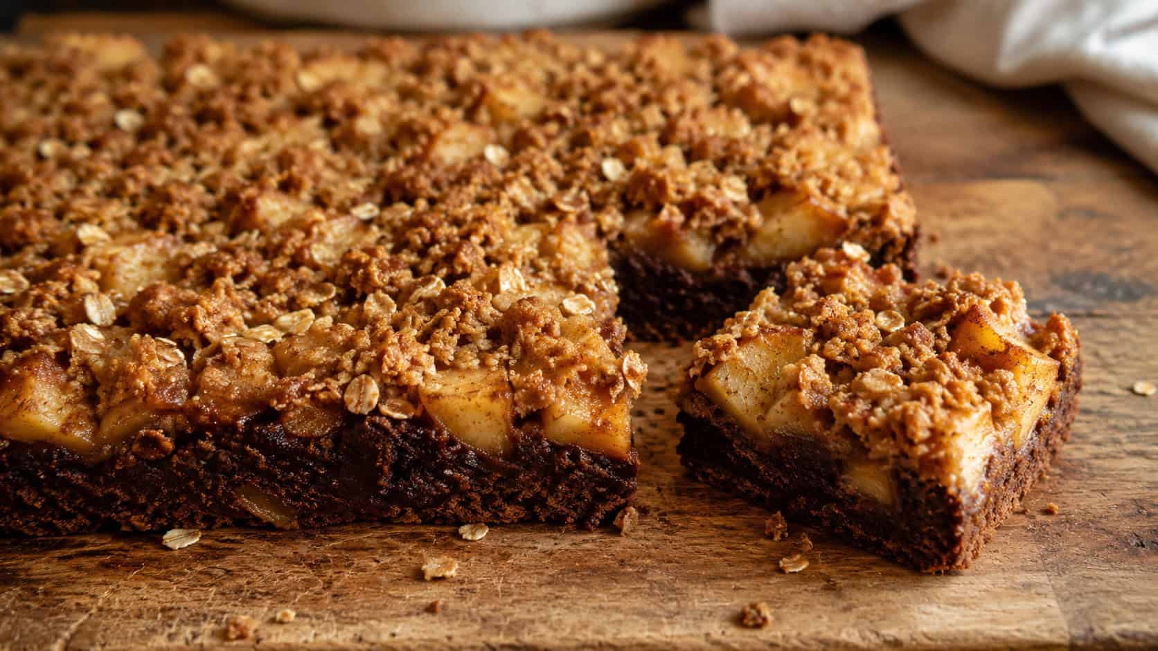 a wide horizontal pan of chocolate brownies topped with cinnamon apples and a golden oat crumble, one piece pulled away, on a rustic wooden board, warm cozy natural light, shallow depth of field, landscape orientation