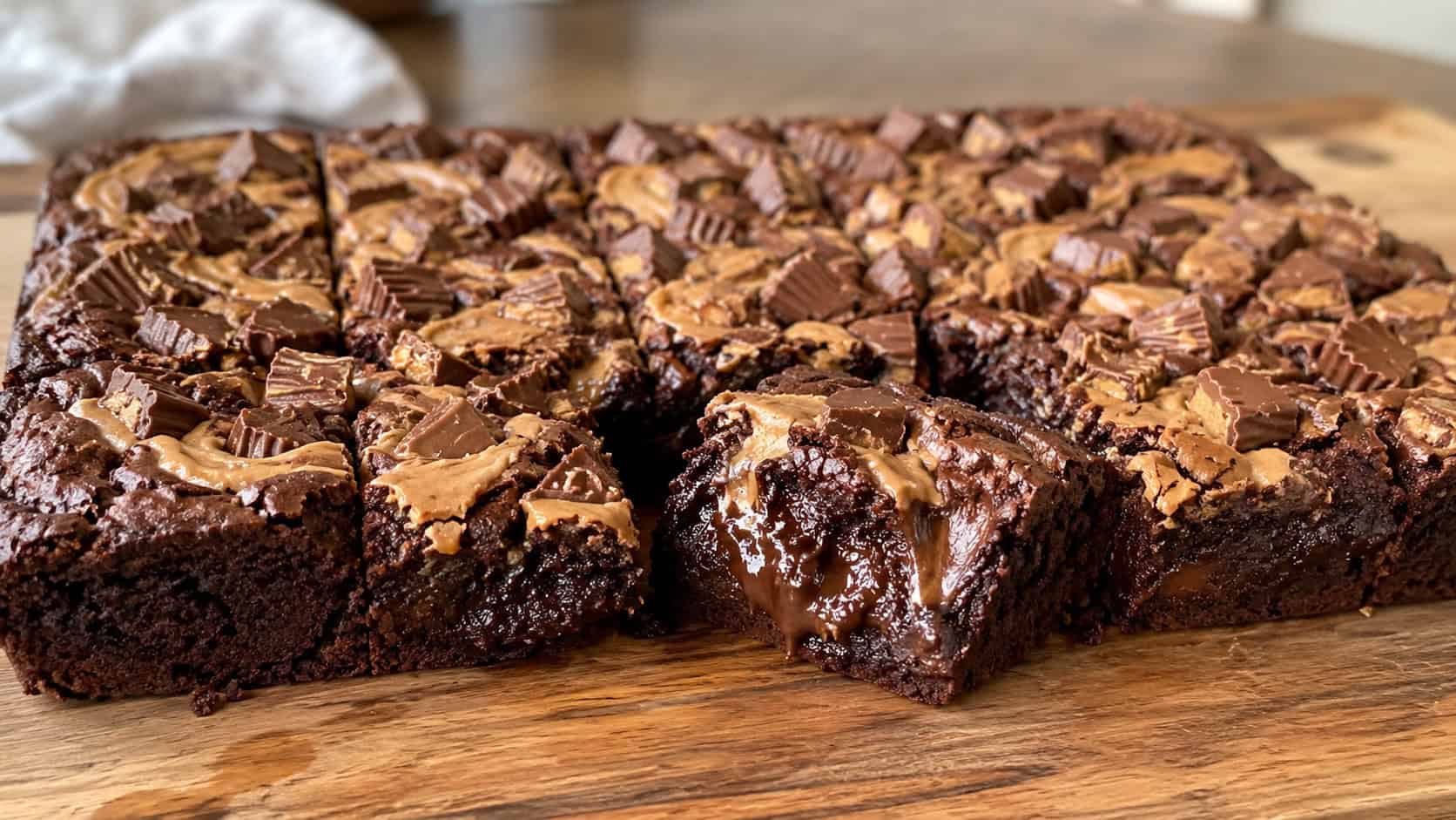 a wide horizontal platter of brownies topped with chopped peanut butter cups and peanut butter swirls, one cut open to show melty chocolate, on a wooden board, natural light, shallow depth of field, landscape orientation