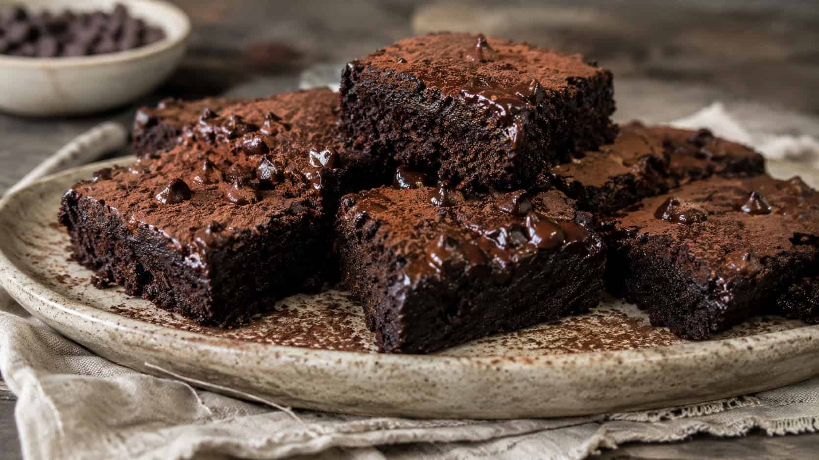 a wide horizontal plate of dark fudgy vegan brownies topped with melting chocolate chips, dusted with cocoa, on a rustic surface with a linen napkin, natural light, shallow depth of field, landscape orientation