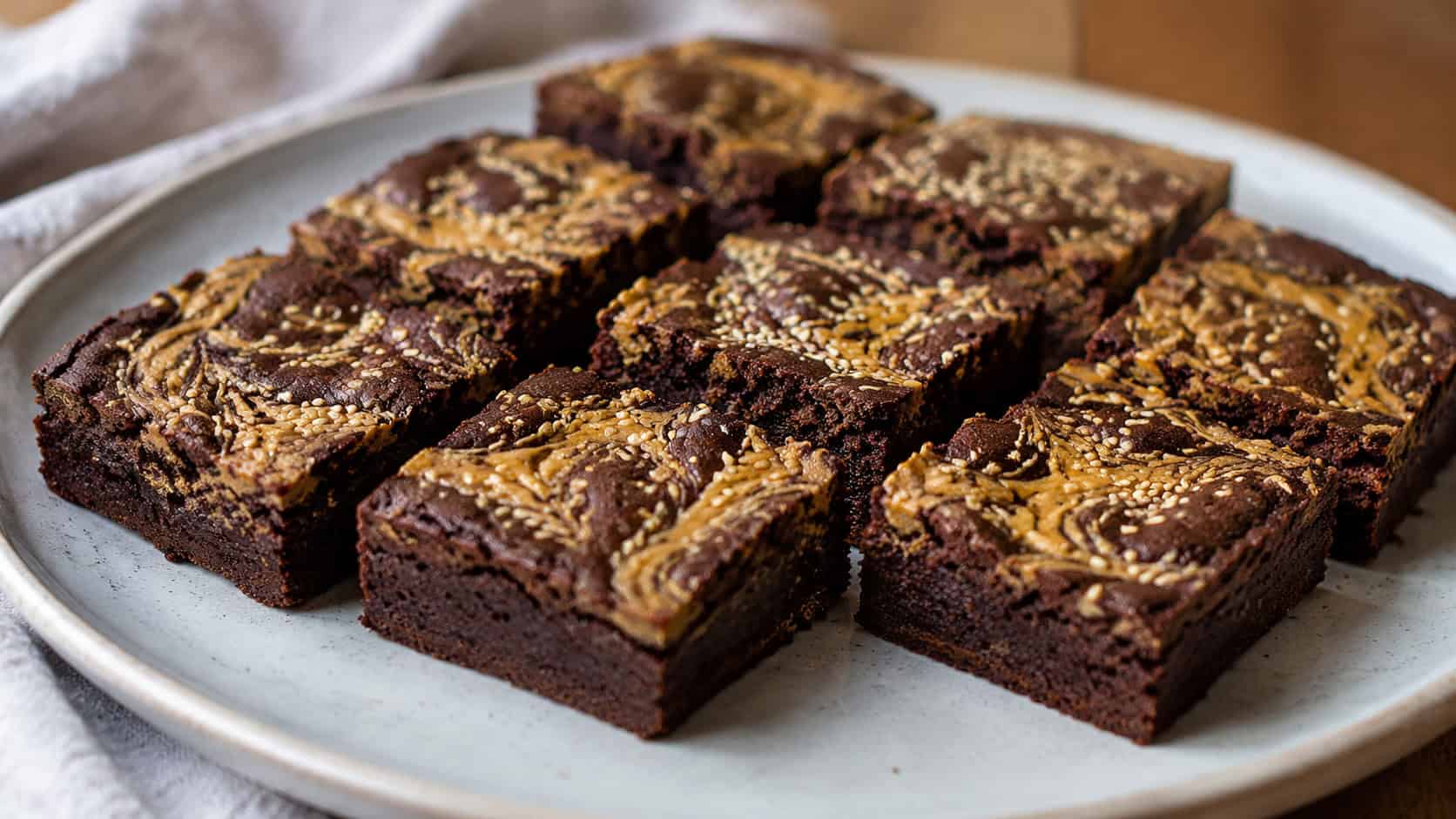 a wide horizontal arrangement of chocolate brownies with golden tahini swirls and sesame seeds on top, on a pale ceramic plate, soft natural light, shallow depth of field, landscape orientation