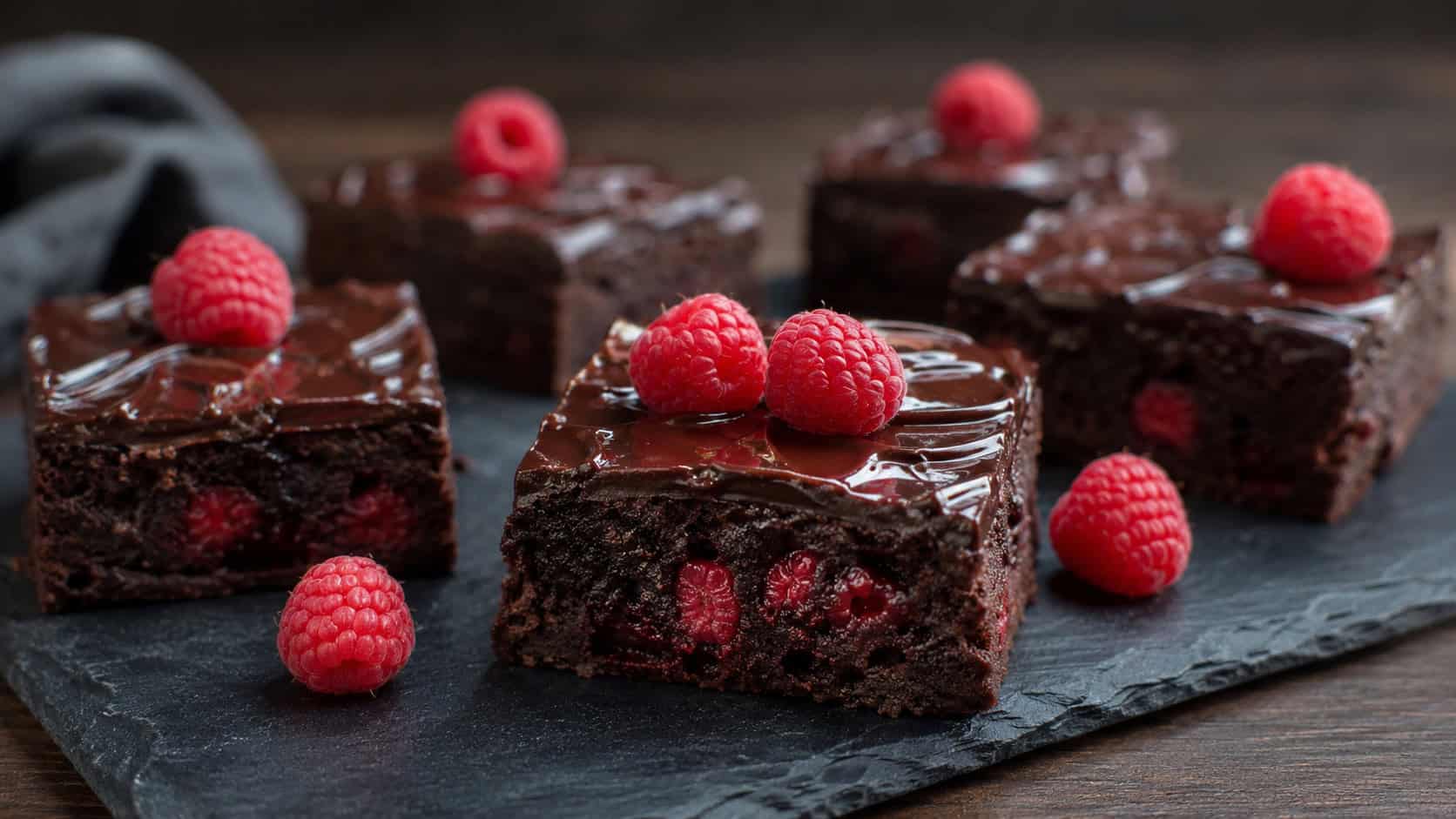 raspberry truffle brownies in a wide horizontal shot, glossy dark ganache top with a few fresh raspberries, rich squares on a slate serving board, moody natural light, shallow depth of field, landscape orientation