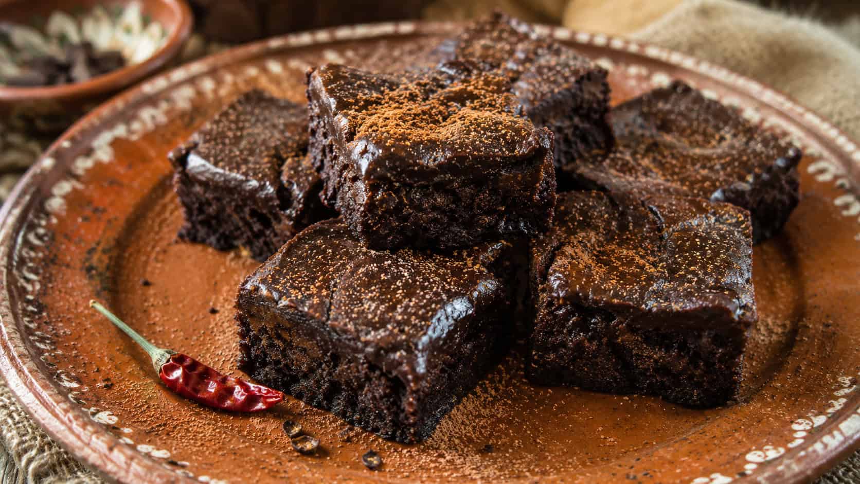 chili chocolate brownies in a wide horizontal shot, dark glossy squares dusted with cinnamon and a tiny chili pepper garnish nearby, on a rustic terracotta plate, warm natural light, shallow depth of field, landscape orientation