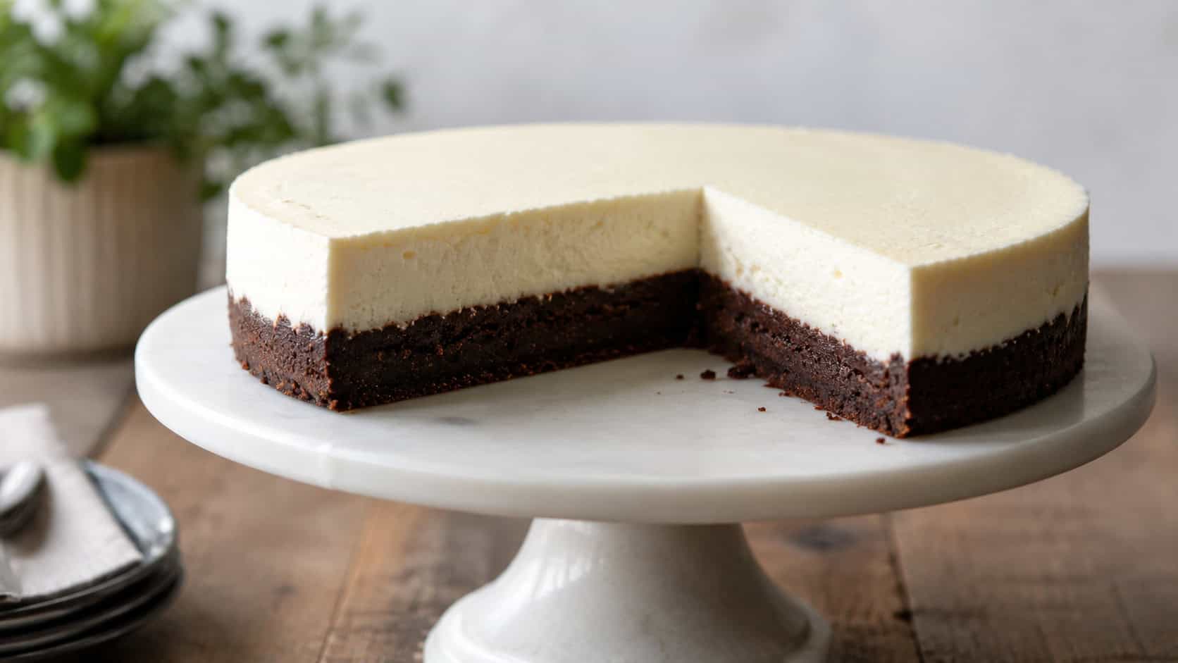 a brownie bottomed cheesecake in a wide horizontal shot, a sliced round cake showing creamy white top over a dark fudgy base, on a footed cake stand, soft natural light, shallow depth of field, landscape orientation