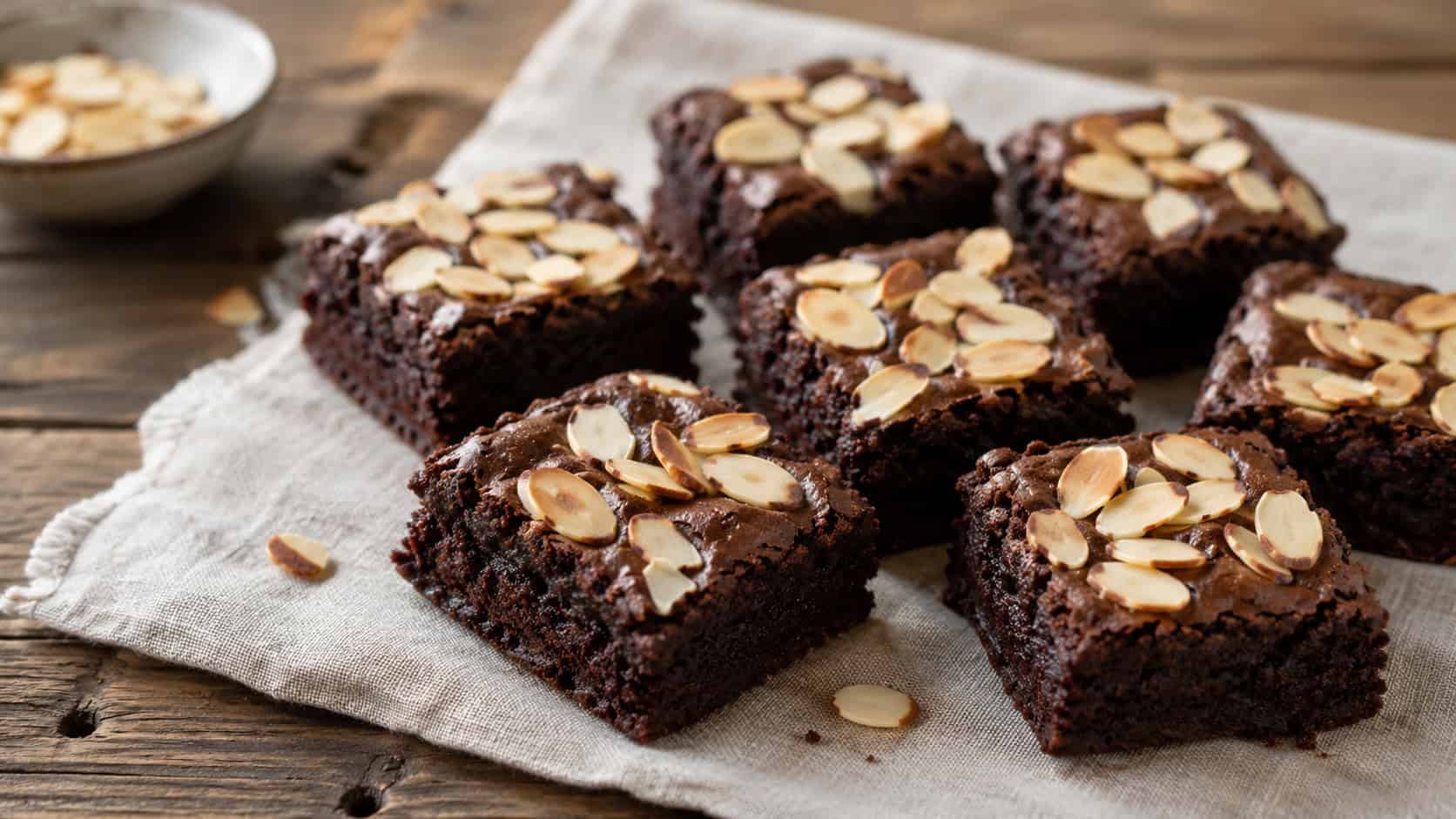 gluten-free almond brownies in a wide horizontal shot, fudgy dark squares topped with toasted sliced almonds, arranged on a linen napkin over a wooden table, natural light, shallow depth of field, landscape orientation