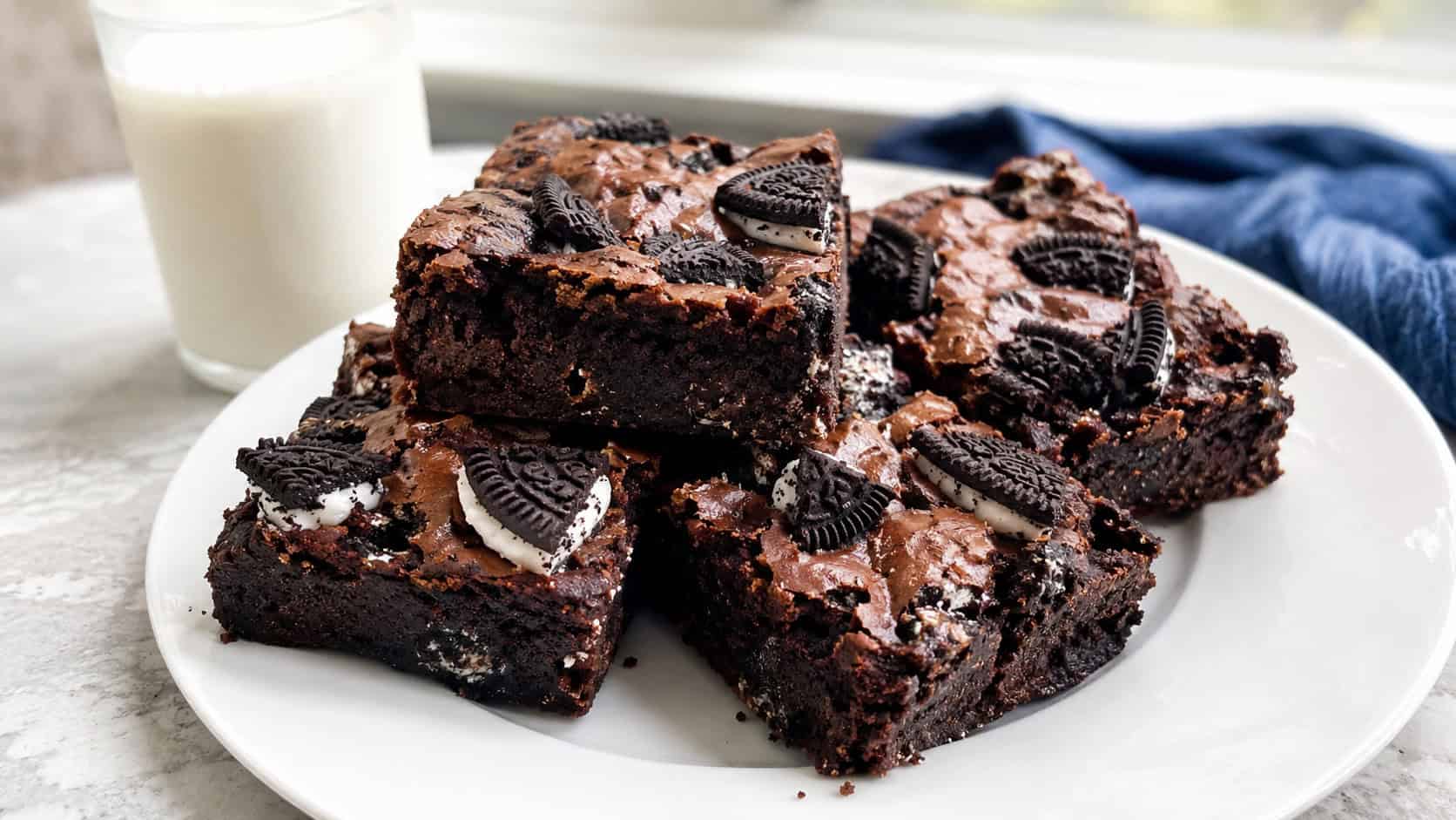 cookies and cream brownies in a wide horizontal shot, fudgy squares loaded with chunks of chocolate sandwich cookies on top, stacked on a white plate with a glass of milk, bright natural light, shallow depth of field, landscape orientation