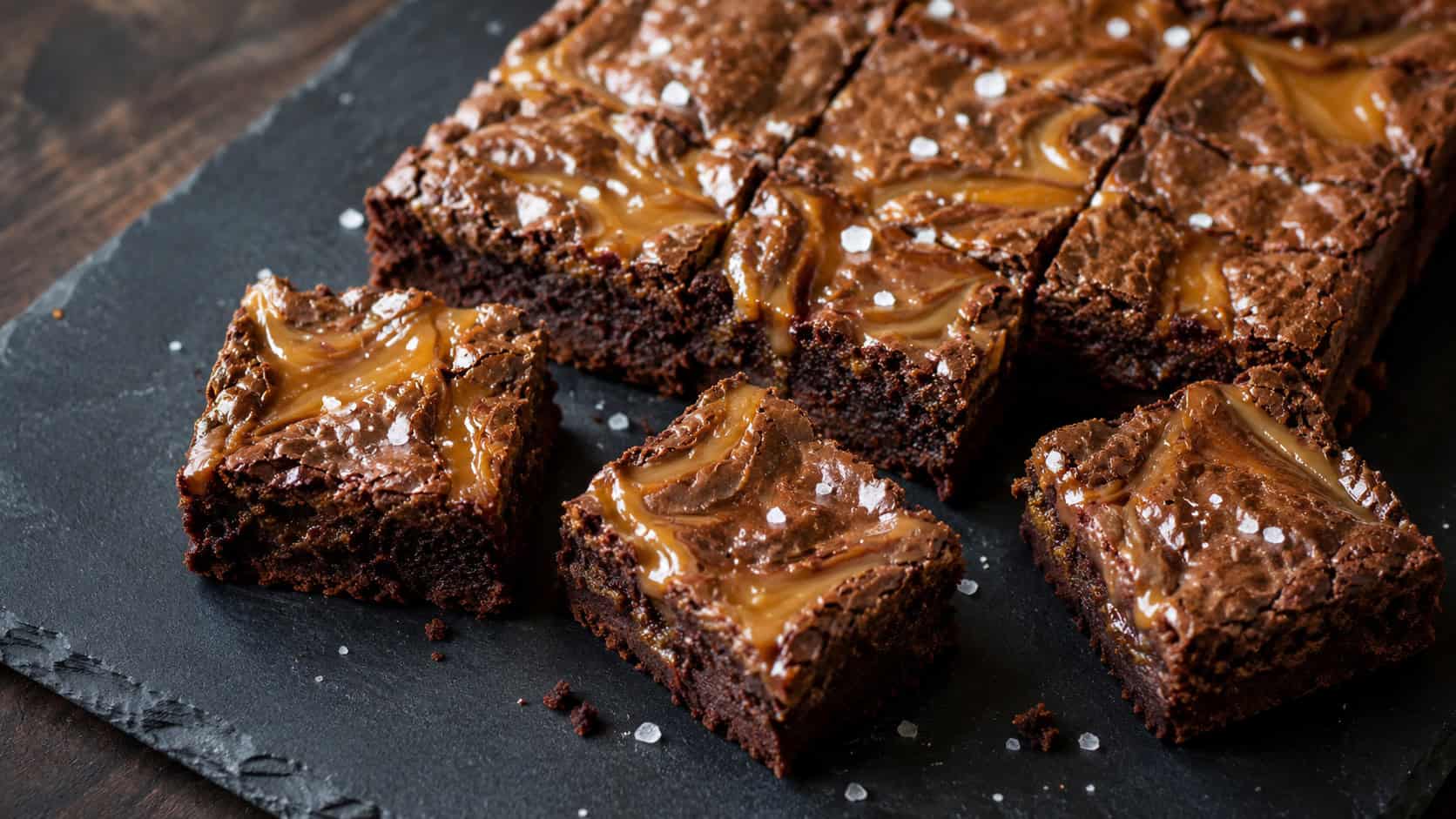salted caramel brownies in a wide horizontal shot, golden caramel swirls glistening on top with flakes of sea salt, a few squares pulled apart on a dark slate board, soft natural light, shallow depth of field, landscape orientation