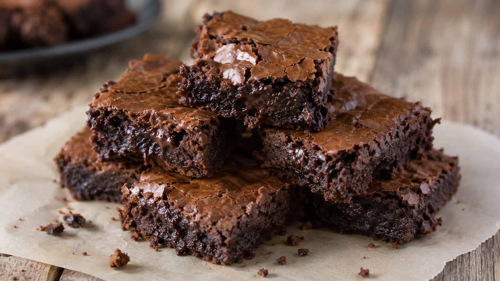 a stack of fudgy chocolate brownies with shiny crackly tops on parchment, wide horizontal shot, gooey interior visible, rustic wooden surface, natural light, shallow depth of field, landscape orientation