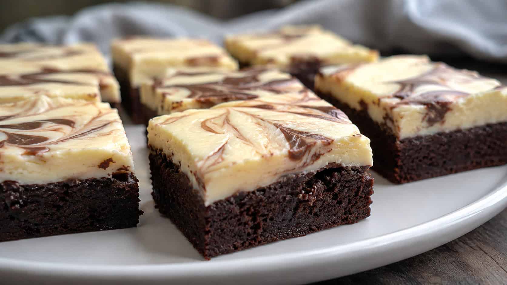 fudgy brownies topped with swirled cream cheese frosting cut into squares on a white plate, wide horizontal shot, thick frosting layer visible, soft natural light, shallow depth of field, landscape orientation