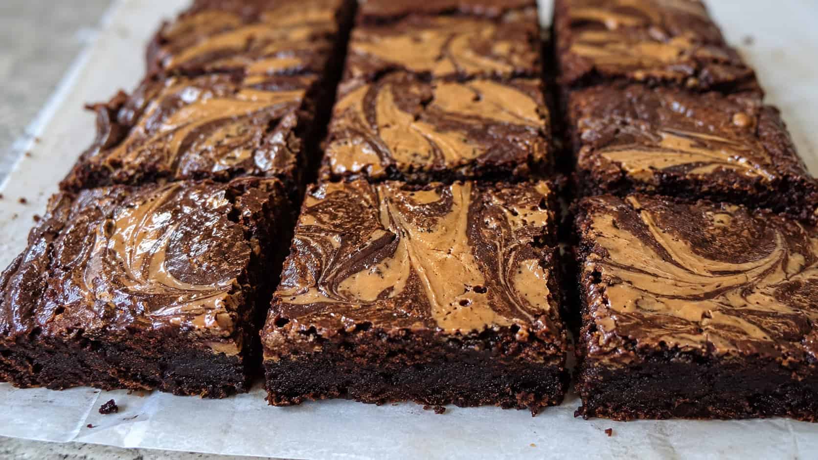fudgy brownies with golden peanut butter swirls on top, cut into squares on parchment, wide horizontal shot, glossy marbled surface, natural light, shallow depth of field, landscape orientation