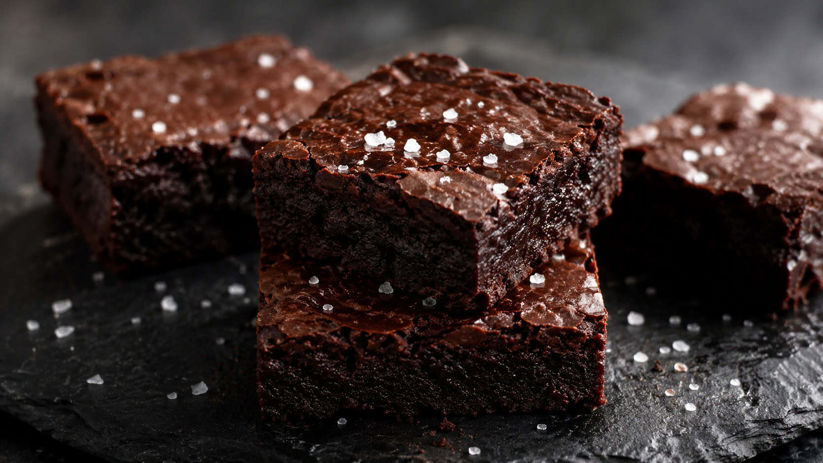 dark chocolate brownies topped with flaky sea salt crystals on a black slate, wide horizontal shot, deep glossy color, dramatic natural light, shallow depth of field, landscape orientation