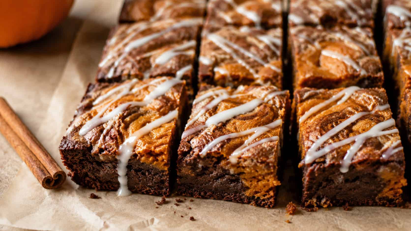 pumpkin brownies with cinnamon swirls and white glaze drizzle cut into squares, in a wide horizontal shot on parchment with cinnamon sticks, natural light, shallow depth of field, landscape orientation