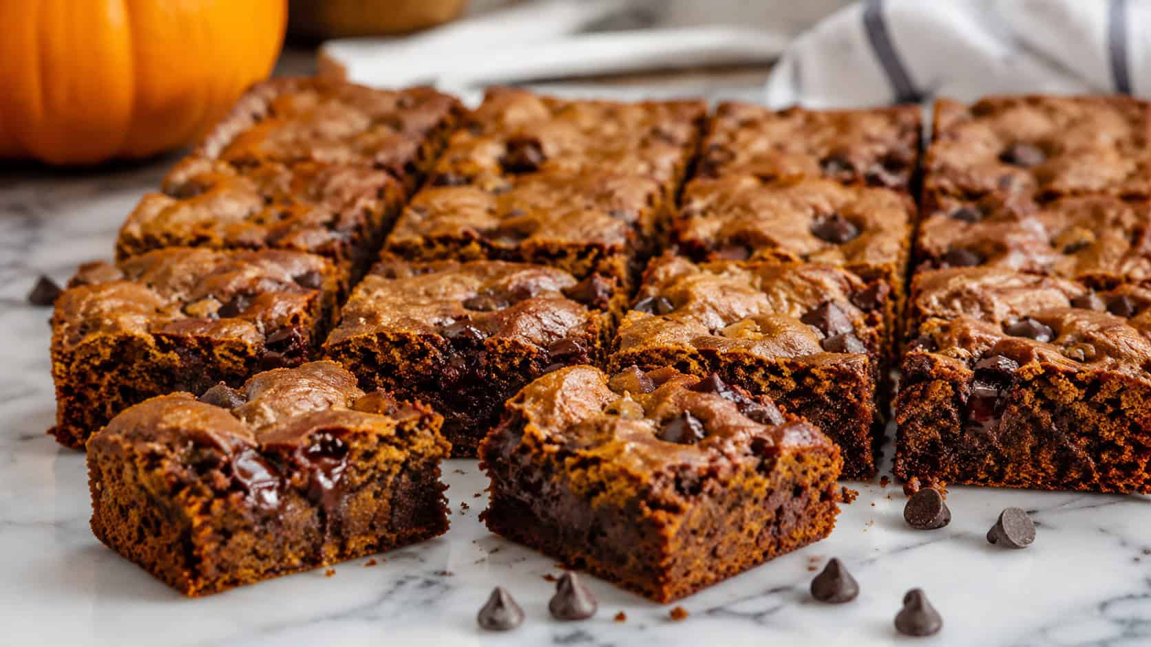 soft pumpkin brownies loaded with melty chocolate chips cut into squares, in a wide horizontal shot on a marble counter with scattered chocolate chips, natural light, shallow depth of field, landscape orientation