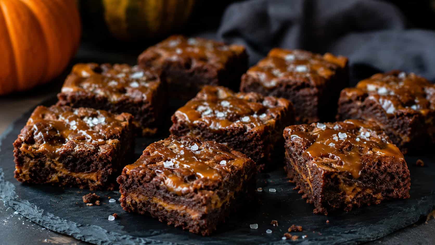 spiced pumpkin brownies with golden caramel ribbons and flaky salt cut into squares, in a wide horizontal shot on a dark slate board, natural light, shallow depth of field, landscape orientation