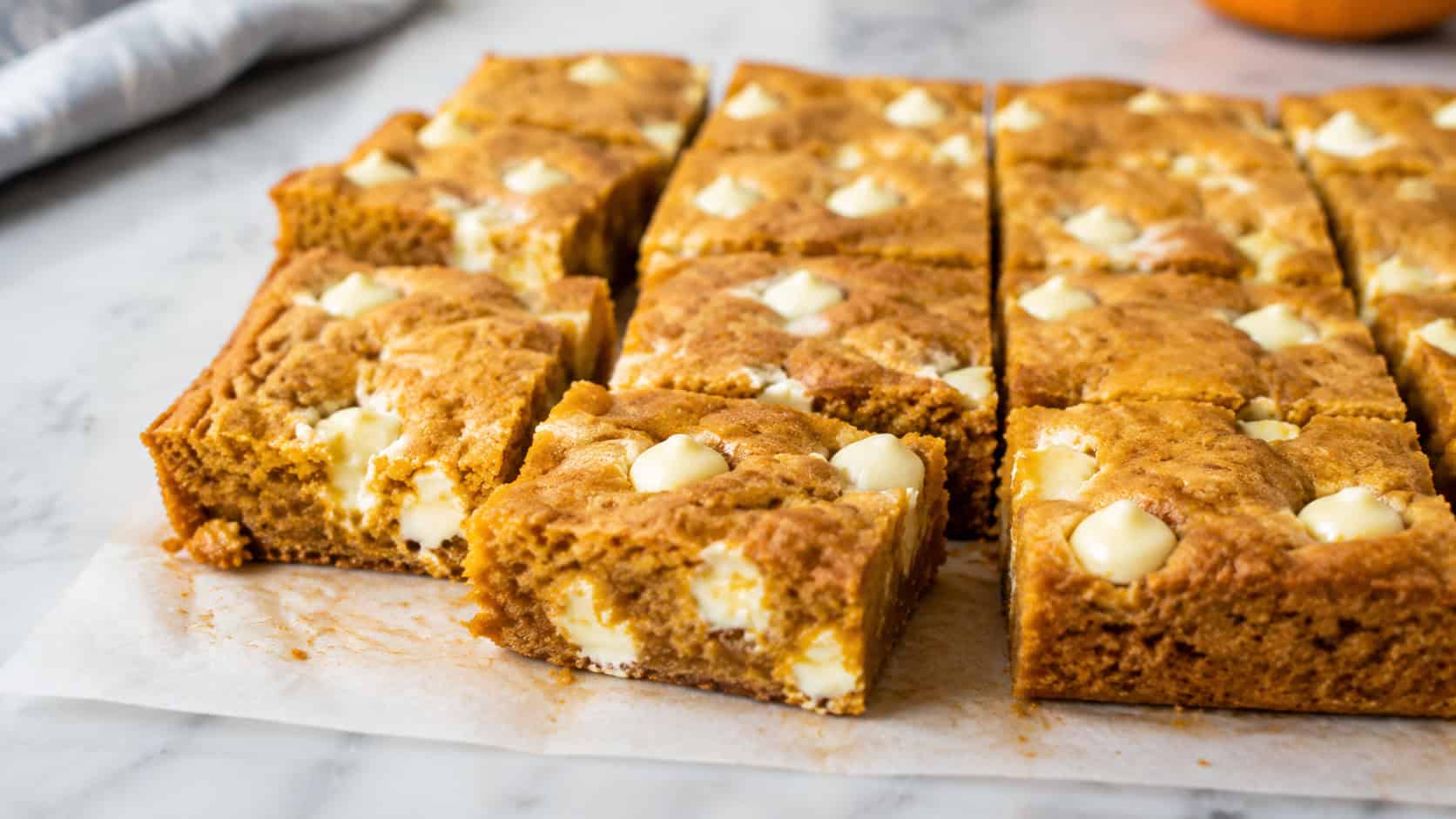 golden pumpkin blondies with melty white chocolate chunks cut into squares, in a wide horizontal shot on parchment over a marble counter, natural light, shallow depth of field, landscape orientation