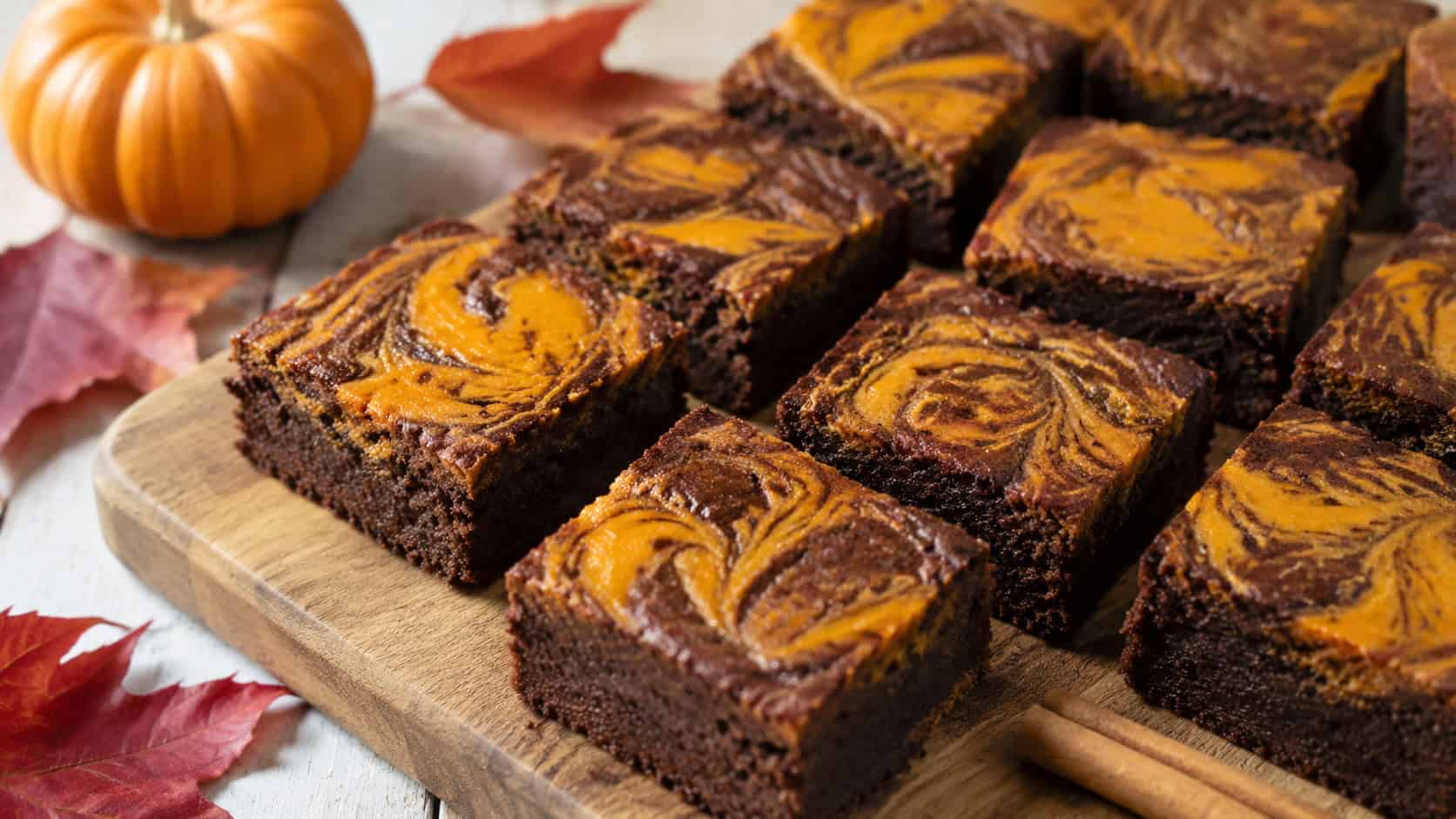 spiced chocolate brownies with a warm orange pumpkin swirl on top in a wide horizontal shot, cut into squares on a wooden board with a cinnamon stick and autumn leaves nearby, natural light, shallow depth of field, landscape orientation