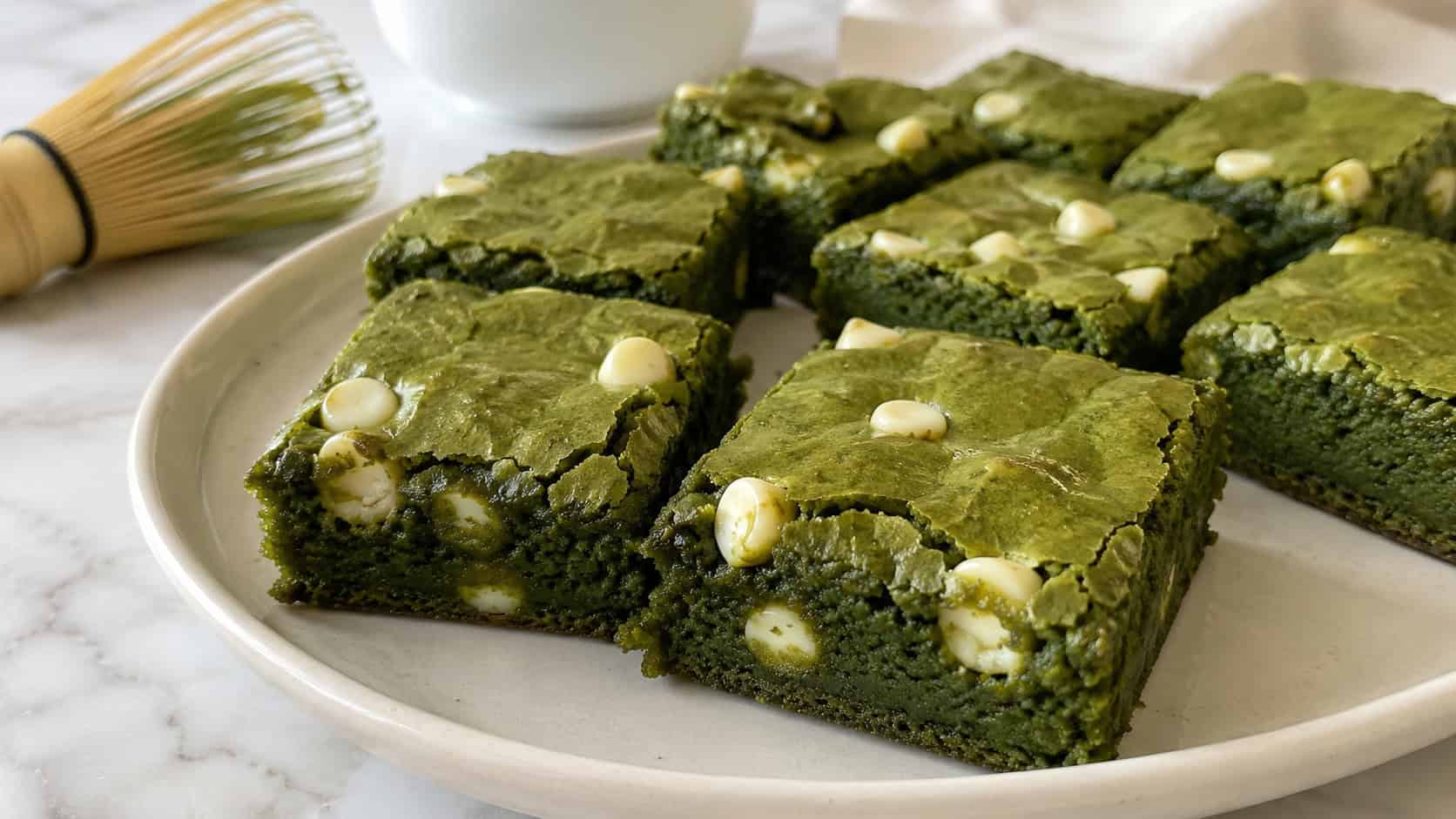 vibrant green matcha brownies with white chocolate chips and a soft chewy edge in a wide horizontal shot, cut into squares on a pale plate beside a small whisk on a bright counter, natural light, shallow depth of field, landscape orientation