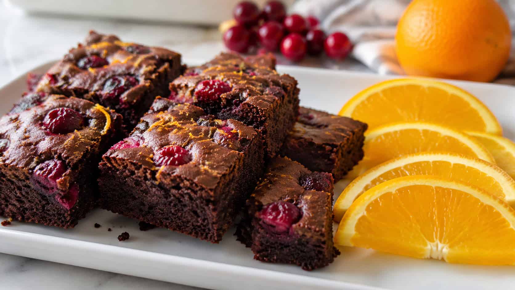 chocolate brownies dotted with ruby cranberries and flecks of orange zest in a wide horizontal shot, cut into squares on a white plate with orange slices beside them, natural light, shallow depth of field, landscape orientation