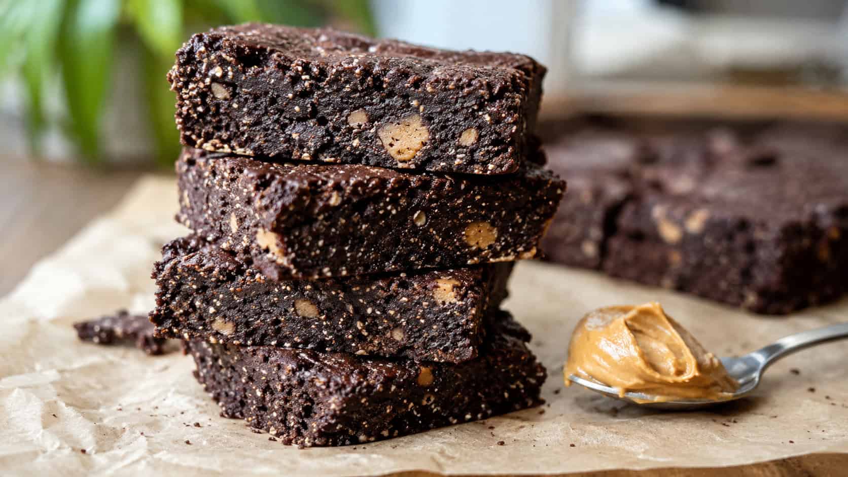 dark peanut butter brownies with a fudgy crumb and visible chia flecks in a wide horizontal shot, stacked on parchment beside a spoonful of peanut butter, natural light, shallow depth of field, landscape orientation