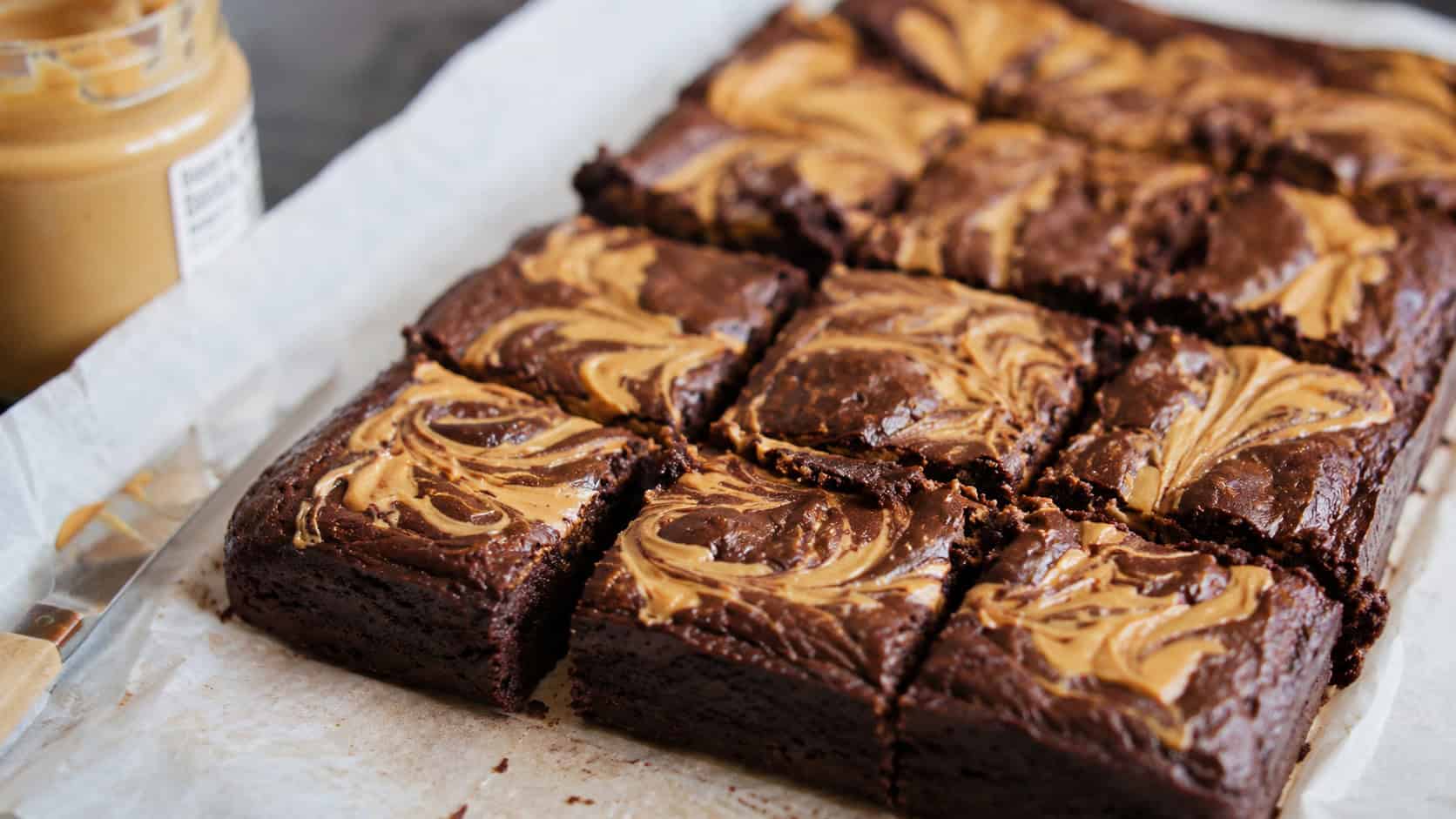 vegan brownies with golden peanut butter swirls marbled across the dark chocolate top in a wide horizontal shot, cut into squares on parchment with a jar of peanut butter nearby, natural light, shallow depth of field, landscape orientation