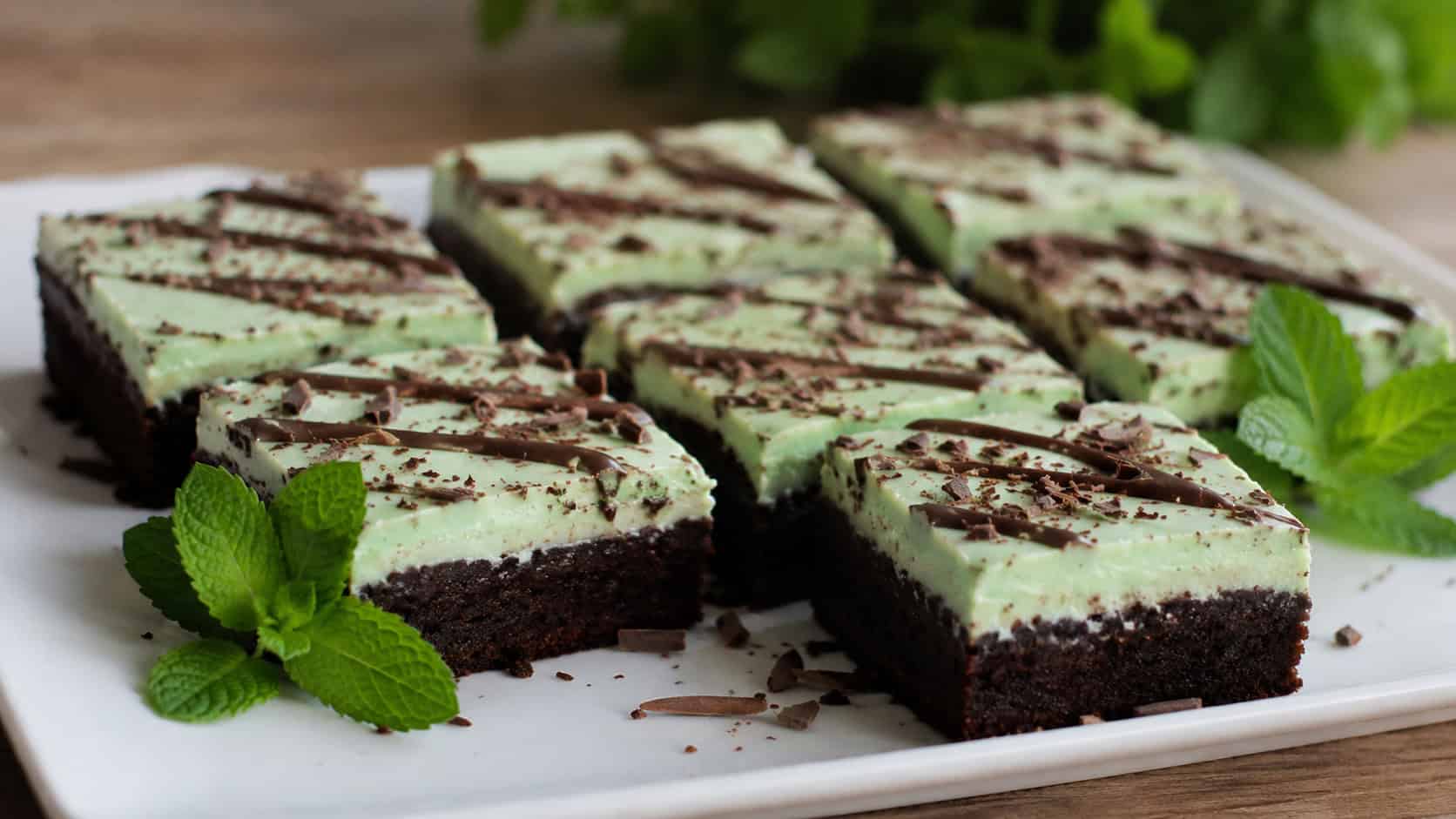 layered brownies with a pale green mint topping and dark chocolate base in a wide horizontal shot, cut into neat squares on a white plate with mint leaves scattered around, natural light, shallow depth of field, landscape orientation