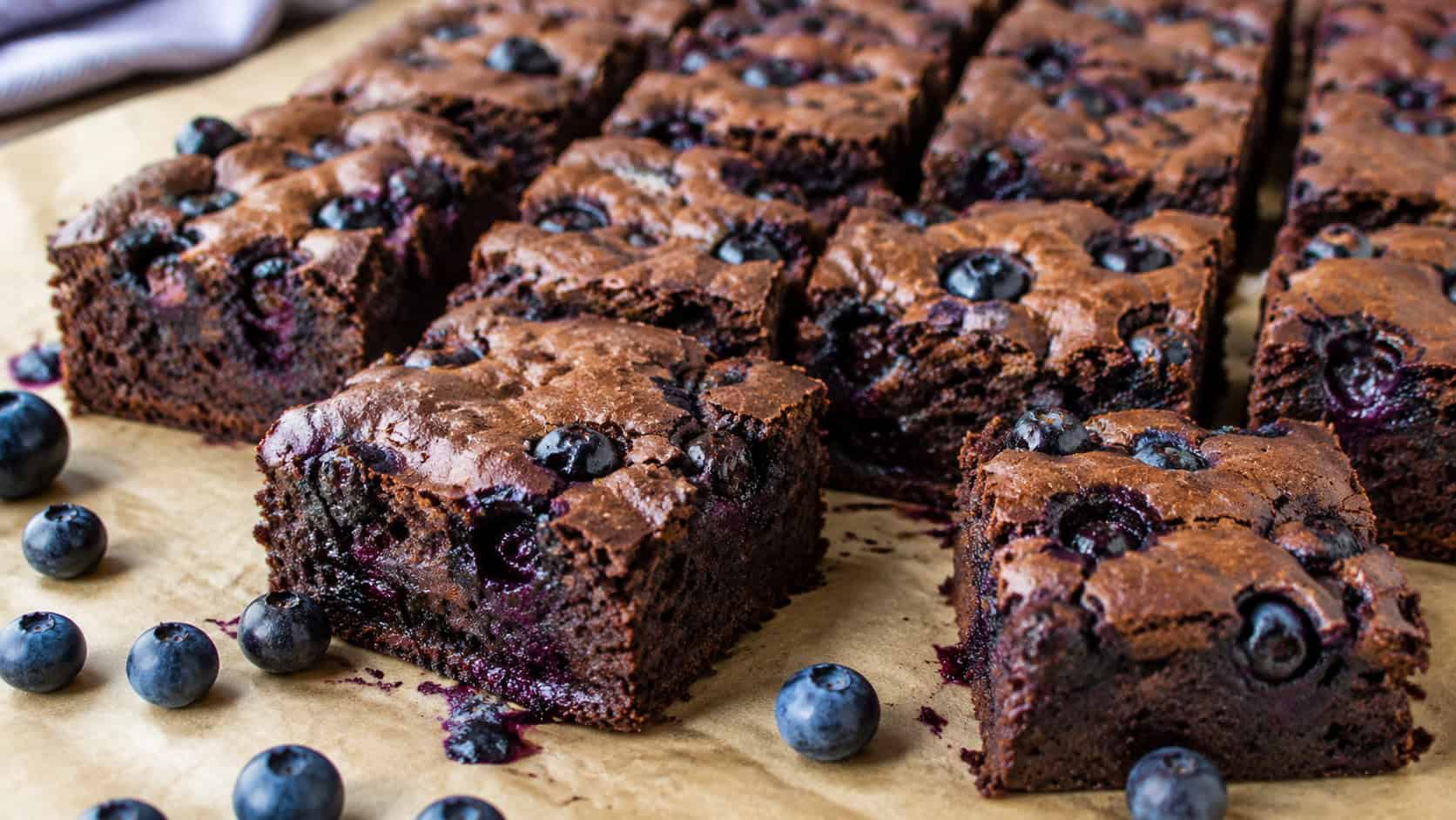 fudgy chocolate brownies studded with glossy blueberries bursting on top in a wide horizontal shot, cut into squares on parchment with fresh blueberries scattered around, natural light, shallow depth of field, landscape orientation
