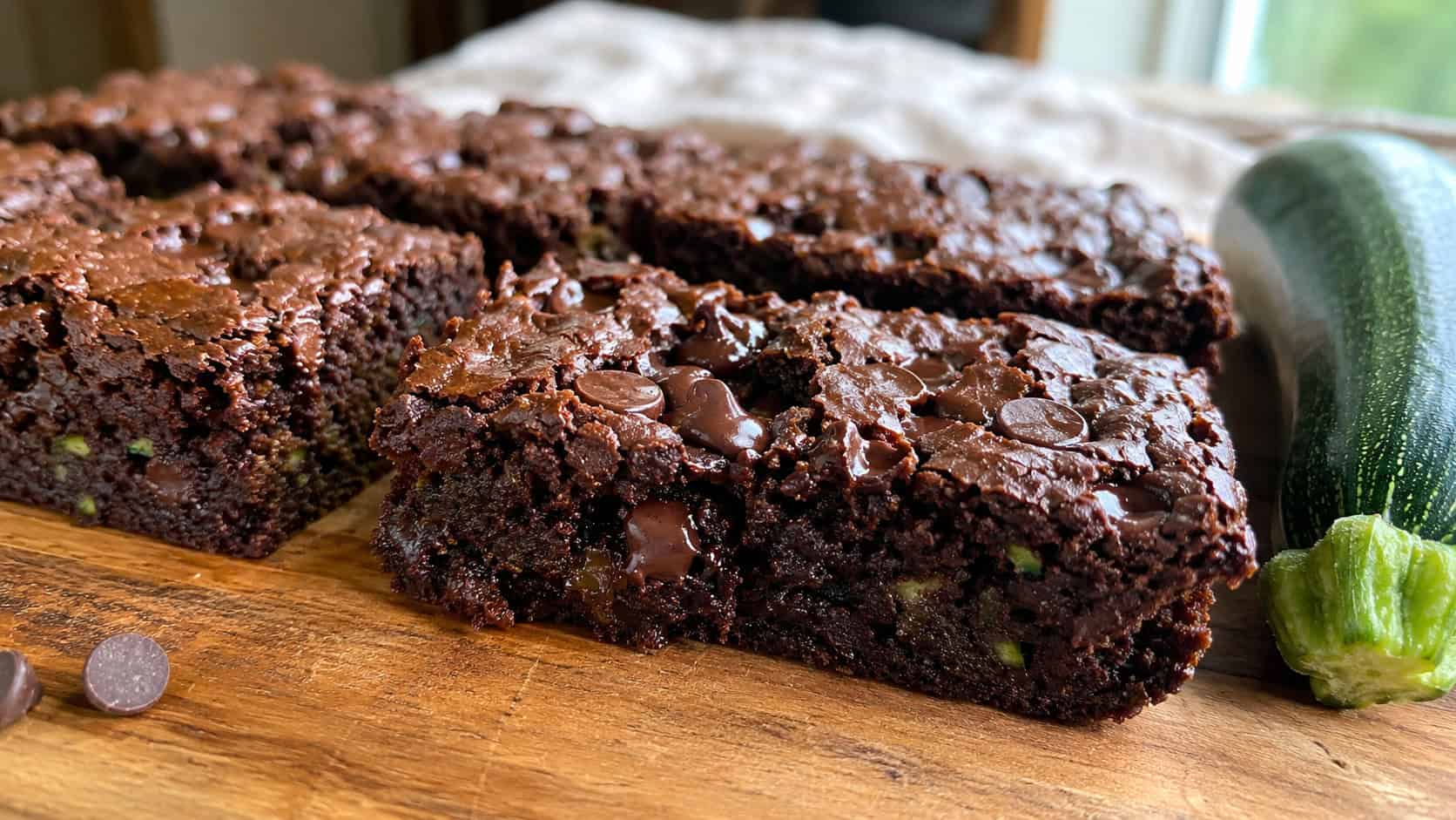 moist chocolate brownies with melty chips and faint green zucchini flecks in a wide horizontal shot, cut into squares on a wooden board with a small zucchini nearby, natural light, shallow depth of field, landscape orientation