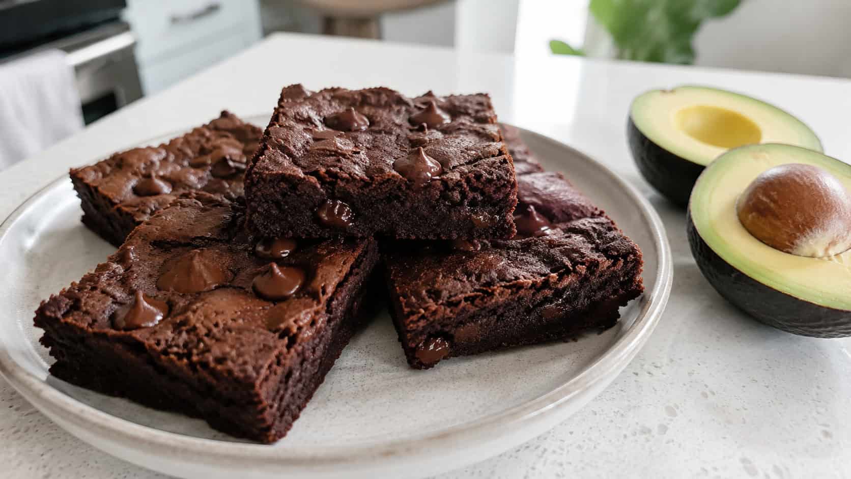 soft fudgy brownies with a velvety dark surface and chocolate chips in a wide horizontal shot, on a ceramic plate beside a halved avocado on a bright kitchen counter, natural light, shallow depth of field, landscape orientation