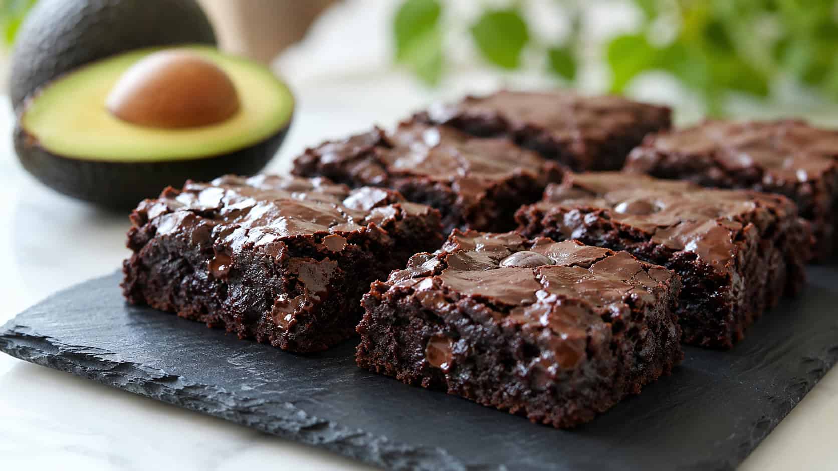 silky dark chocolate brownies on a slate board in a wide horizontal composition, glossy fudgy surface with melted chips, a halved avocado softly blurred in the background, bright natural light, shallow depth of field, landscape orientation