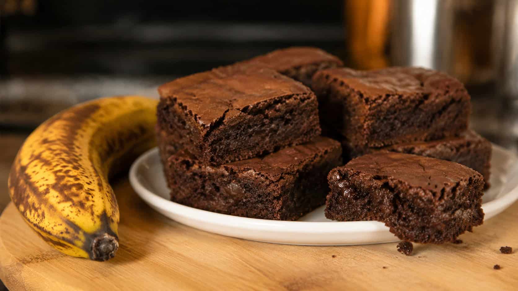 soft chocolate brownies on a wooden board in a wide horizontal shot, one square broken to show the tender crumb, a spotty ripe banana resting beside the plate, warm kitchen light, shallow depth of field, landscape orientation