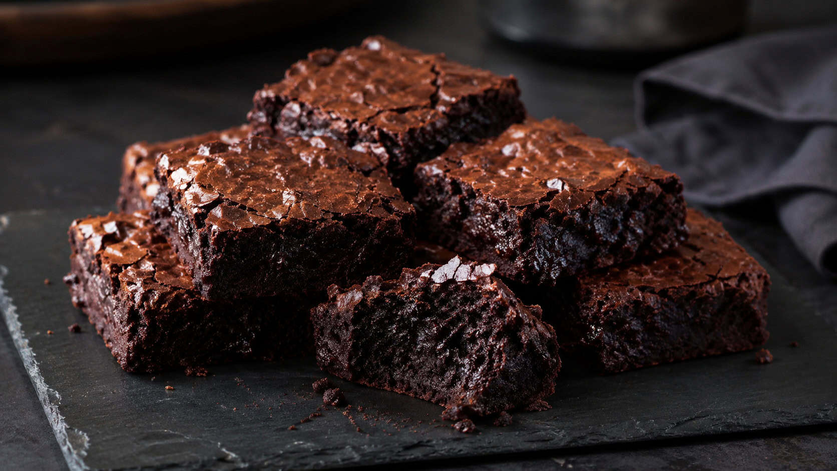 intensely dark fudgy brownies on a black slate plate in a wide horizontal shot, glossy crackled tops catching the light, a square broken to show the dense interior, moody dark table, soft natural light, shallow depth of field, landscape orientation