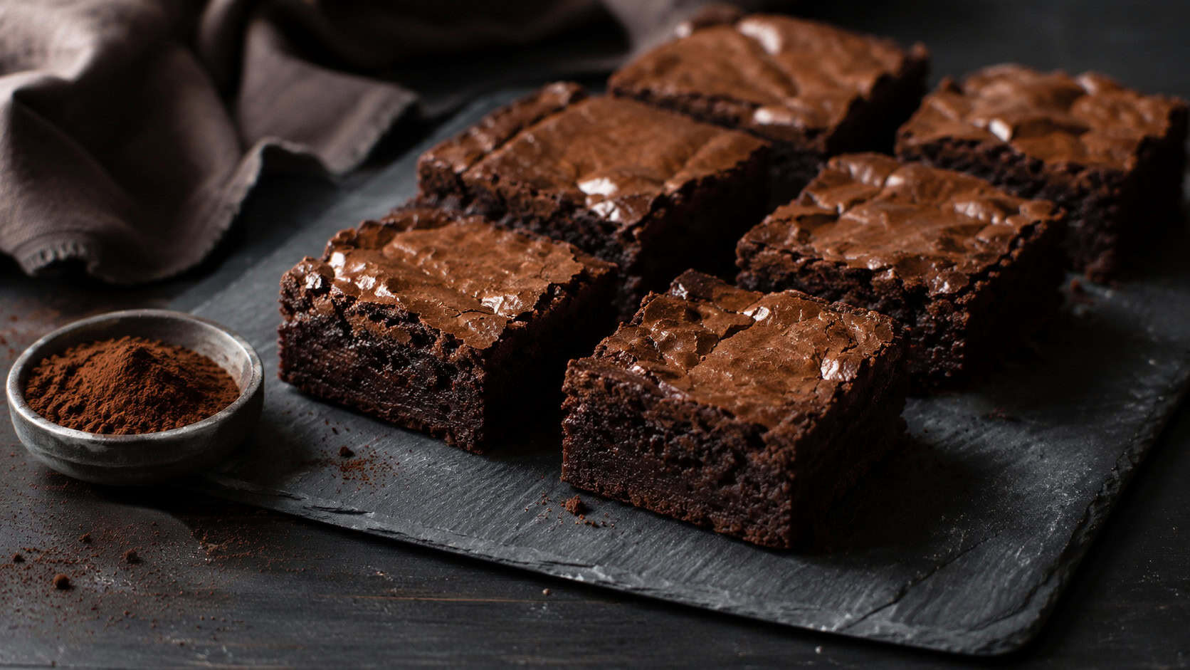 dark vegan fudge brownies arranged on a slate board in a wide horizontal composition, glossy tops with a soft crackle, a small bowl of cocoa powder beside them, linen napkin on a moody dark table, natural light, shallow depth of field, landscape orientation