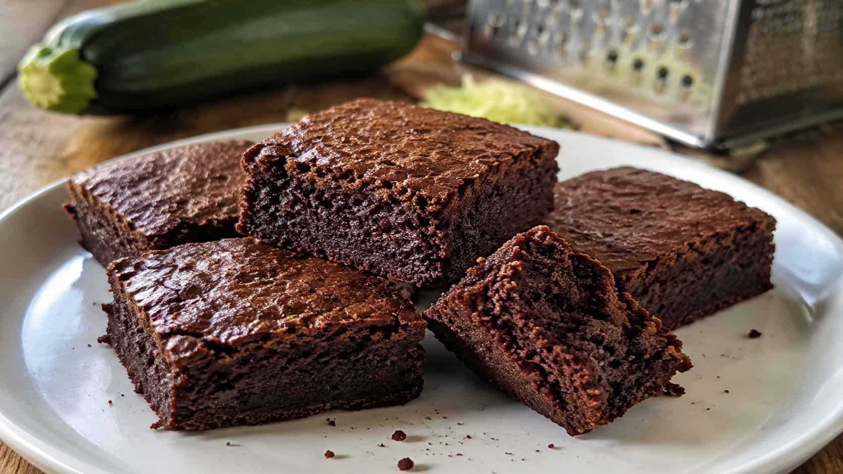 moist chocolate brownies on a white plate in a wide horizontal shot, one square broken to show the dense crumb, a fresh zucchini and grater softly blurred in the background on a wooden table, natural light, shallow depth of field, landscape orientation