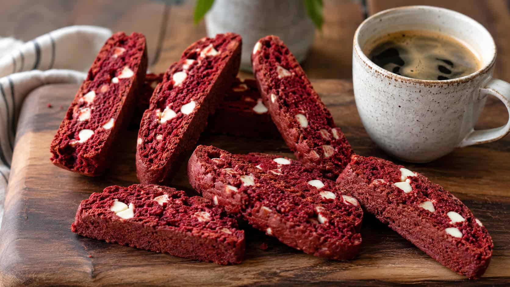 a wide horizontal arrangement of red velvet biscotti slices with white chocolate chips, standing and lying on a wooden board beside a coffee cup, natural light, shallow depth of field, landscape orientation