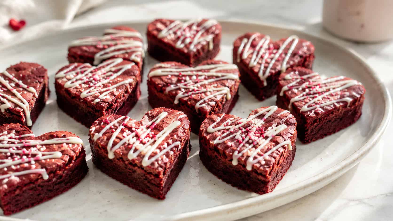 a wide horizontal platter of heart-shaped red velvet brownies drizzled with white frosting and a few sprinkles, on a pale ceramic plate, bright natural light, shallow depth of field, landscape orientation