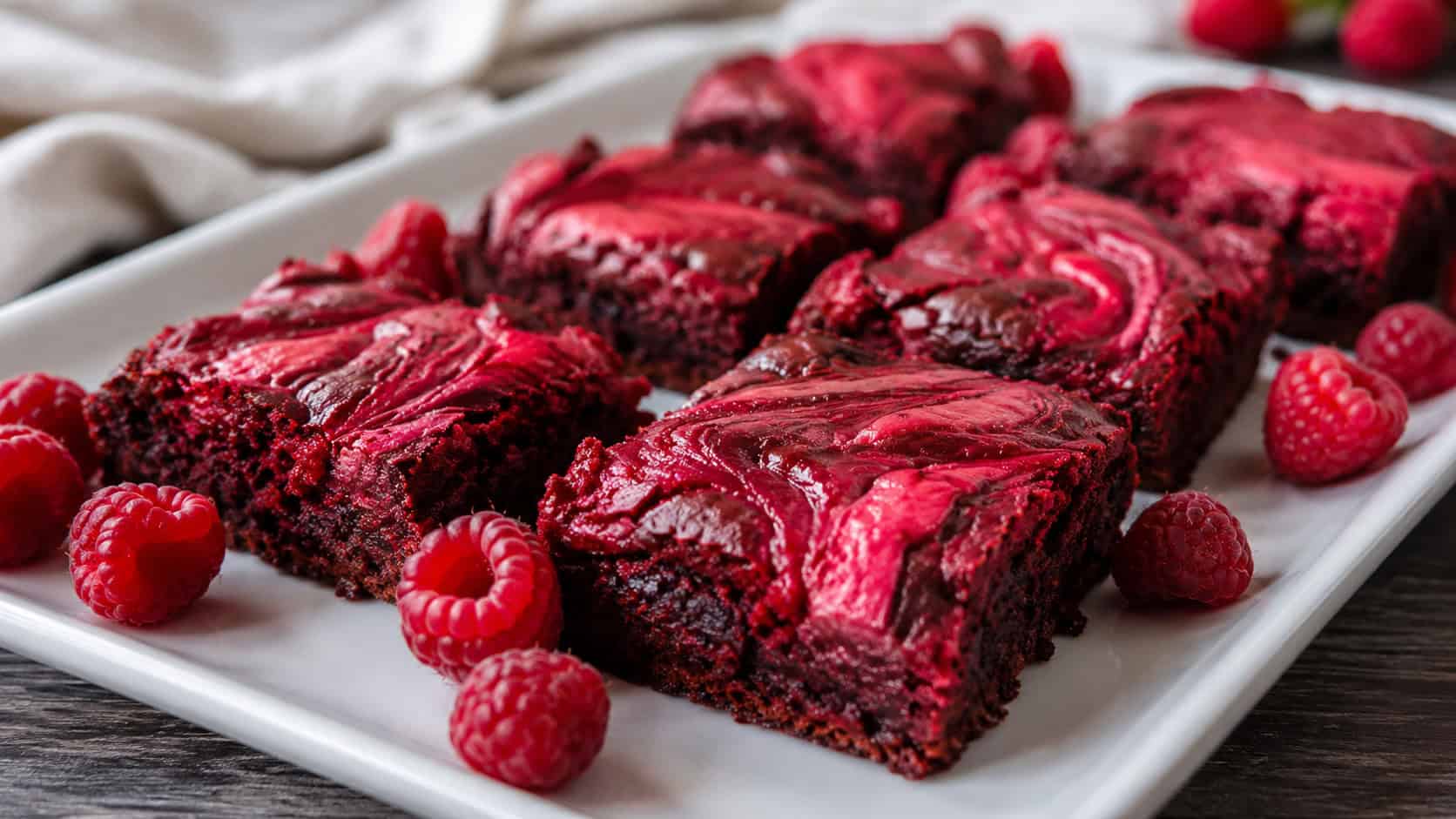 a wide horizontal platter of red velvet brownies with bright raspberry ripples swirled on top, fresh raspberries scattered around, on a white plate, natural light, shallow depth of field, landscape orientation