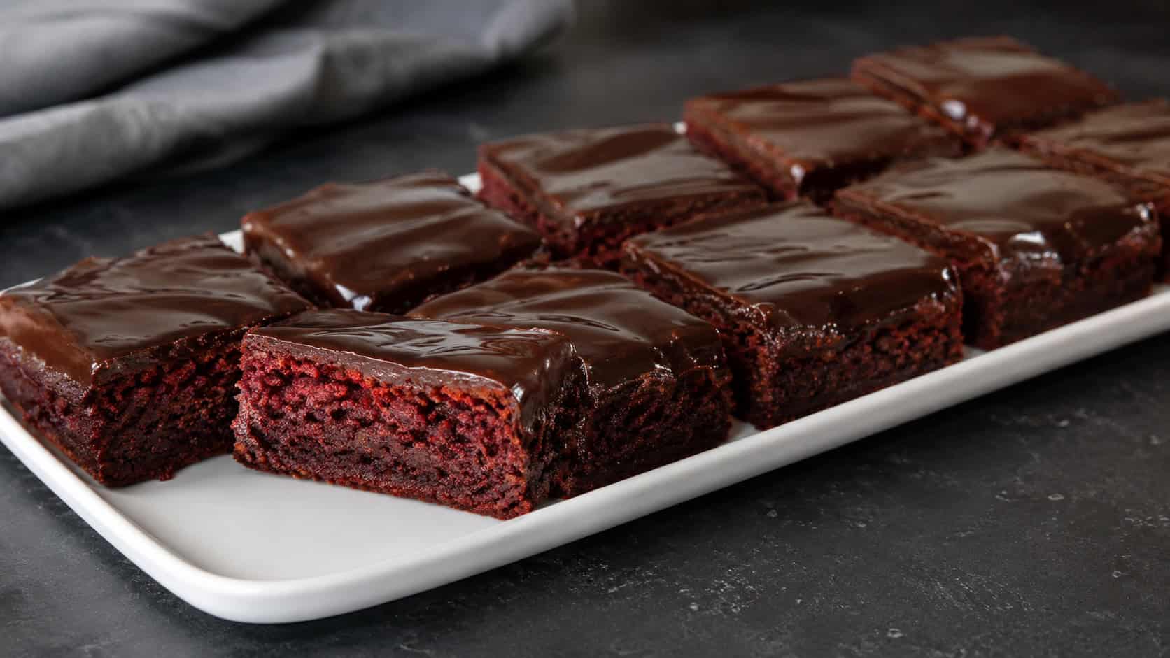a wide horizontal platter of red velvet brownies topped with a glossy dark chocolate ganache glaze, one cut to show the red base, on a dark surface, soft natural light, shallow depth of field, landscape orientation