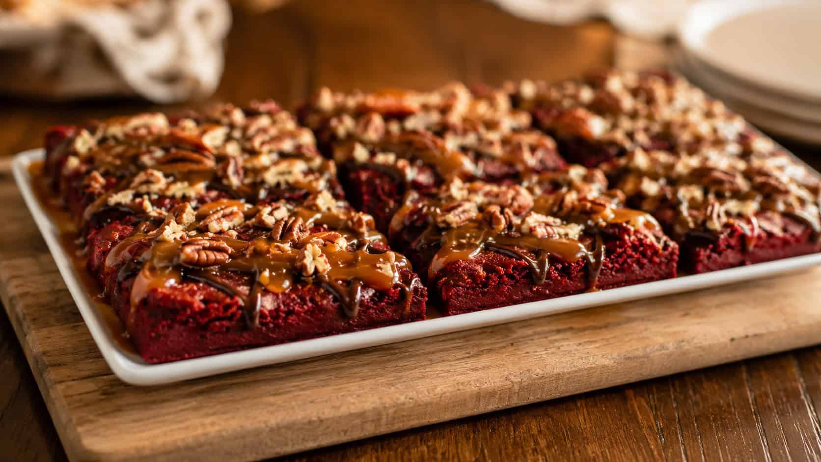 a wide horizontal platter of red velvet turtle brownies drizzled with caramel and chocolate and topped with toasted pecans, on a wooden board, warm natural light, shallow depth of field, landscape orientation