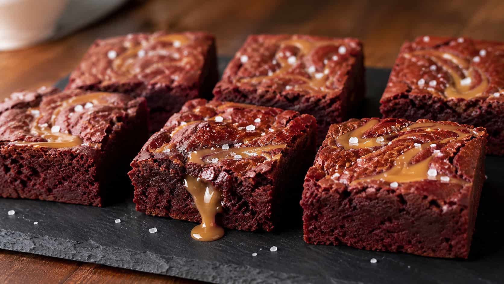 a wide horizontal arrangement of red velvet brownies with golden caramel swirls and flaky salt on top, one piece with caramel oozing out, on a dark slate board, warm natural light, shallow depth of field, landscape orientation