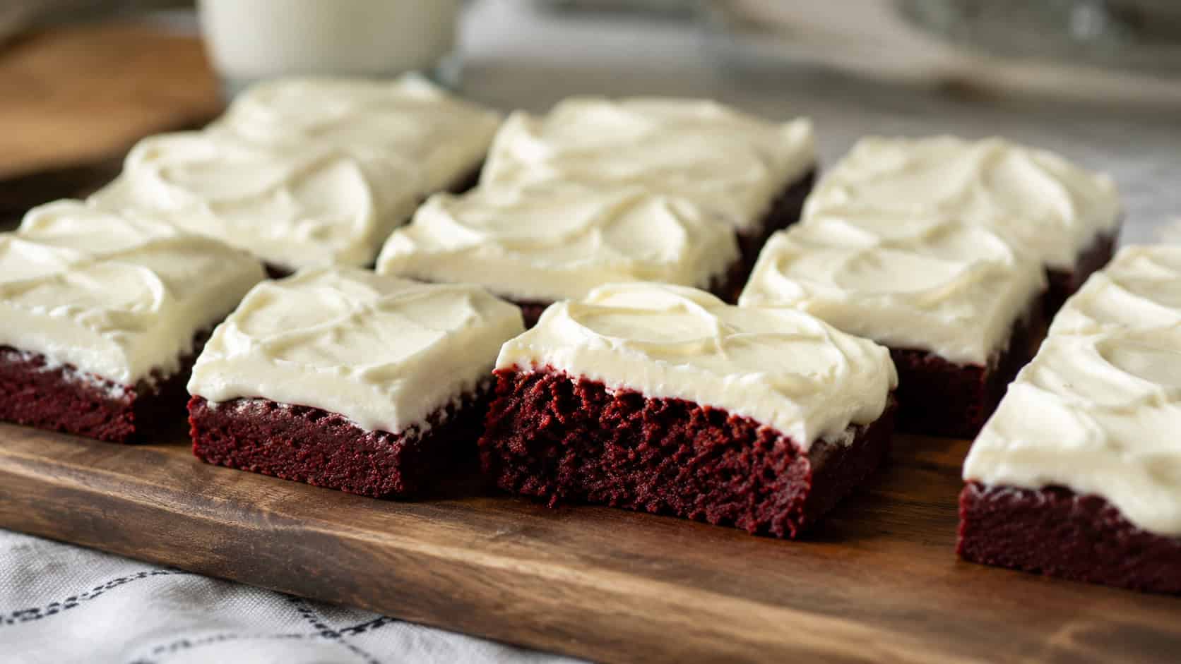 a wide horizontal arrangement of red velvet brownies topped with a thick layer of swirled white cream cheese frosting, one piece cut to show the red base, on a wooden board, soft natural light, shallow depth of field, landscape orientation